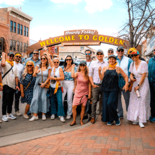 A group of Golden History Tours participants poses in front of Golden, Colorado's street-spanning "Welcome to Golden" sign.